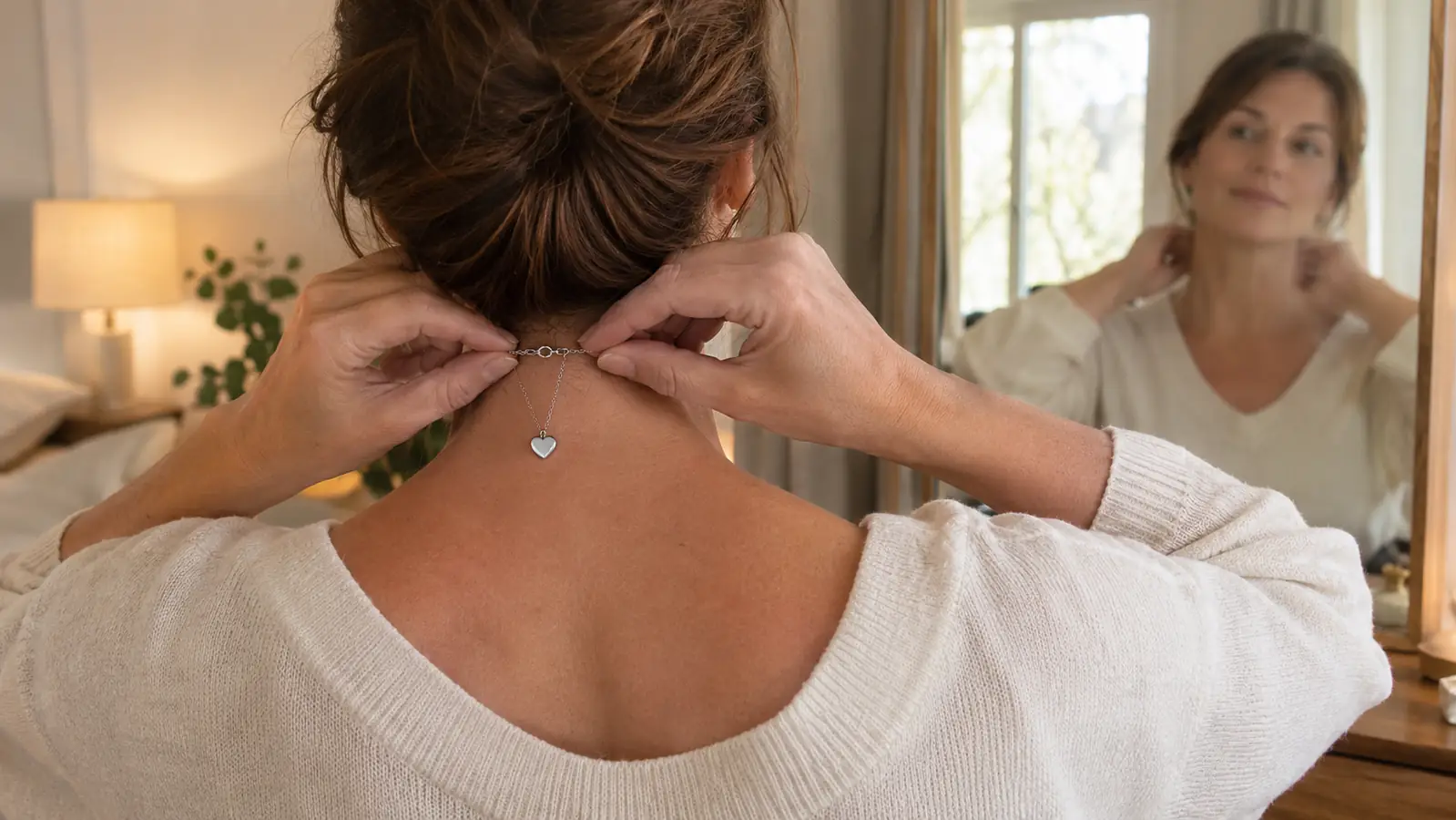 A woman fastening the clasp of a heart necklace at the back of her neck in front of a mirror