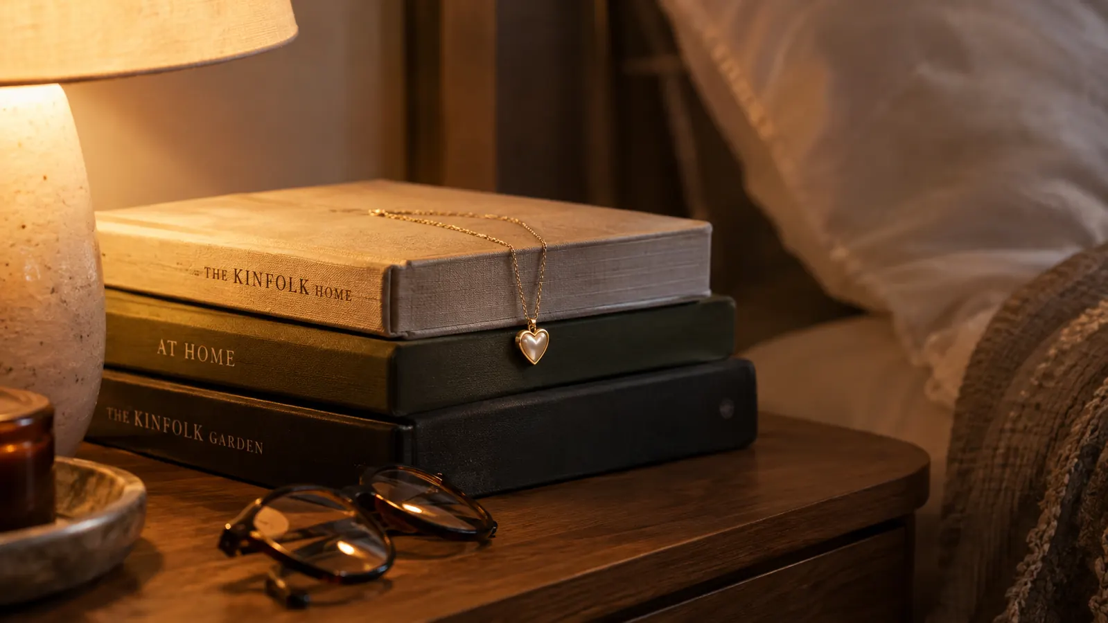 A heart necklace draped over a small stack of hardcover books on a nightstand beside a reading lamp