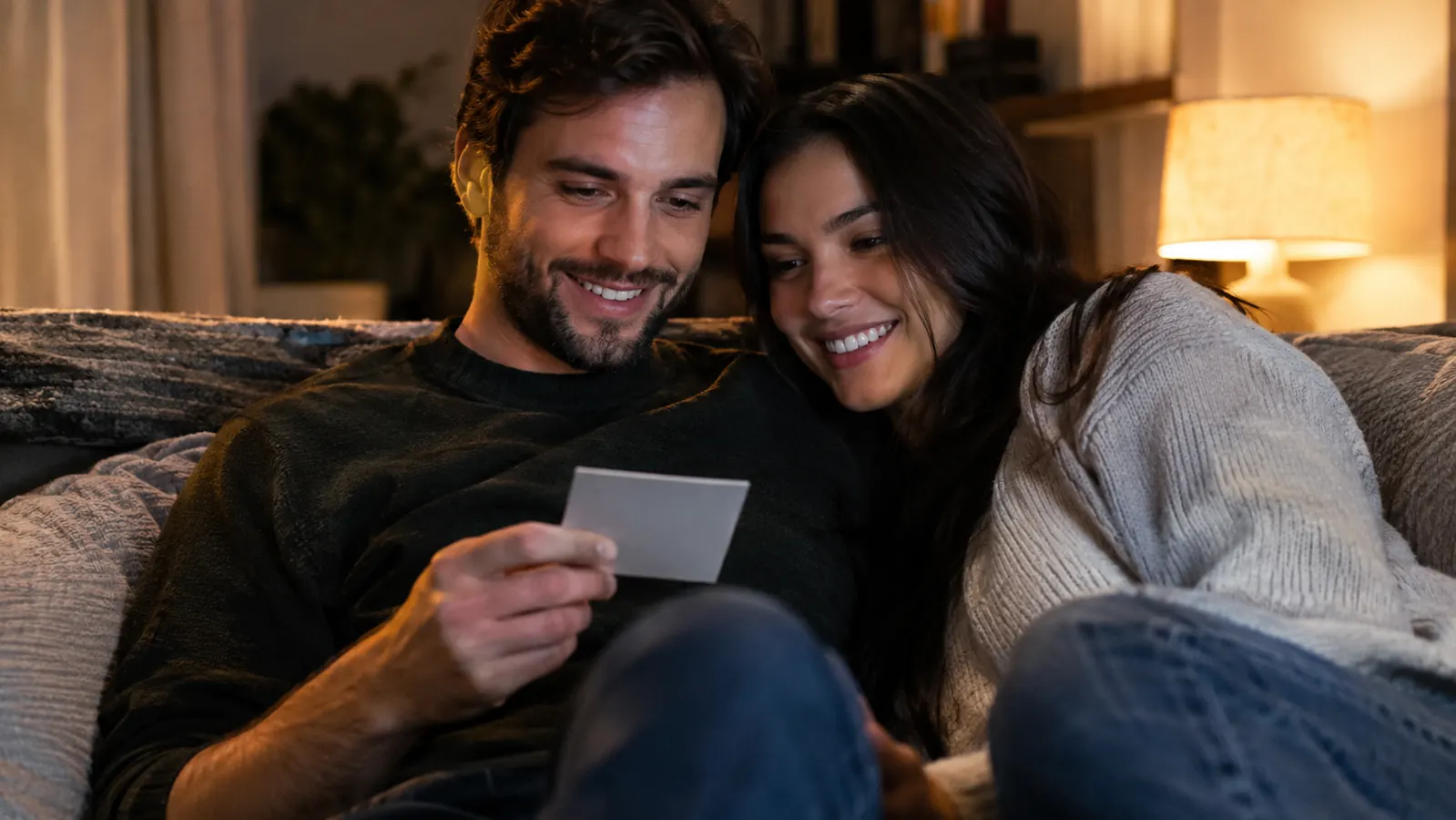 A couple sitting close together on a couch, one holding a small card and smiling, the other leaning in to look, with a lamp glowing in the evening